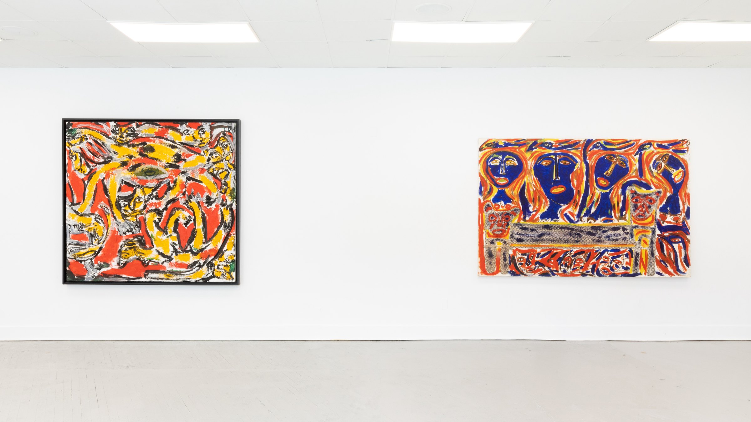 Two paintings by Thornton Dial hang far apart on a long white gallery wall. The left one, framed in black, is a mass of red, yellow and white loops with faces and a large eye worked into it; the right one, unframed, shows four blue faces above a patterned bench against red and orange.