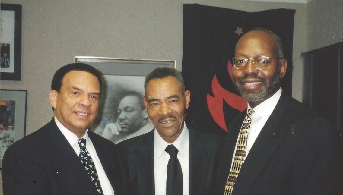 Three men stand together for a photograph in an office: Ambassador Andrew Young at the left, Thornton Dial at the centre in a dark suit and tie, and Jerry Thomas at the right in glasses. On the wall behind them hang a framed photograph of Martin Luther King Jr. and a dark banner.
