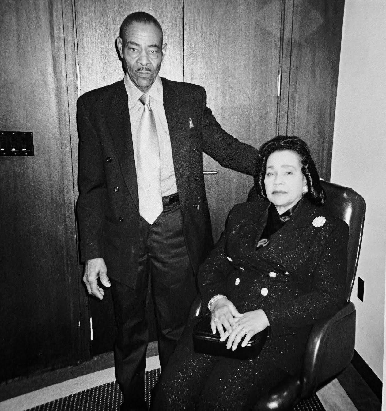 A black-and-white photograph of Thornton Dial standing in a suit and pale tie beside Coretta Scott King, who is seated in a leather armchair in a dark jacket with a brooch, her hands resting on a clutch bag. Both look toward the camera; the room is panelled in dark wood.