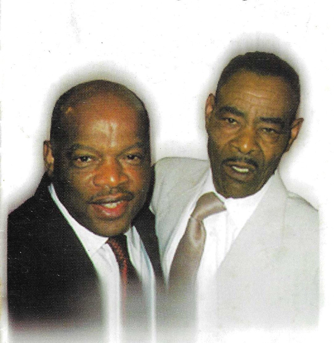 A close photograph of Thornton Dial, in a pale suit at the right, standing shoulder to shoulder with Congressman John Lewis, in a dark suit at the left. Both are smiling at the camera against a white background.