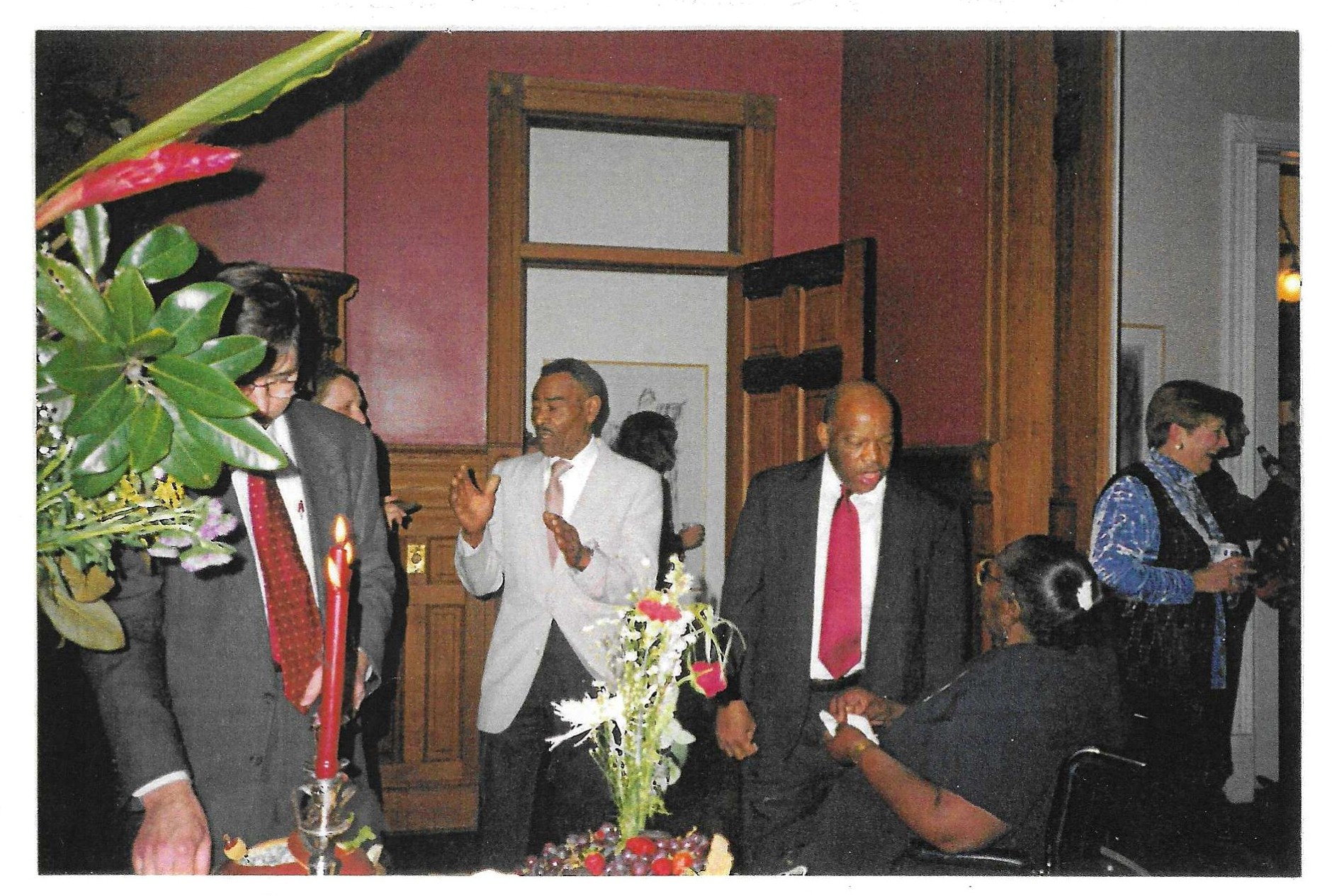 Thornton Dial, in a pale suit at the centre, speaks with his hands raised at a reception in a red-walled room. Congressman John Lewis stands to his right in a dark suit and red tie, and other guests stand around a table set with flowers, fruit and lit candles.