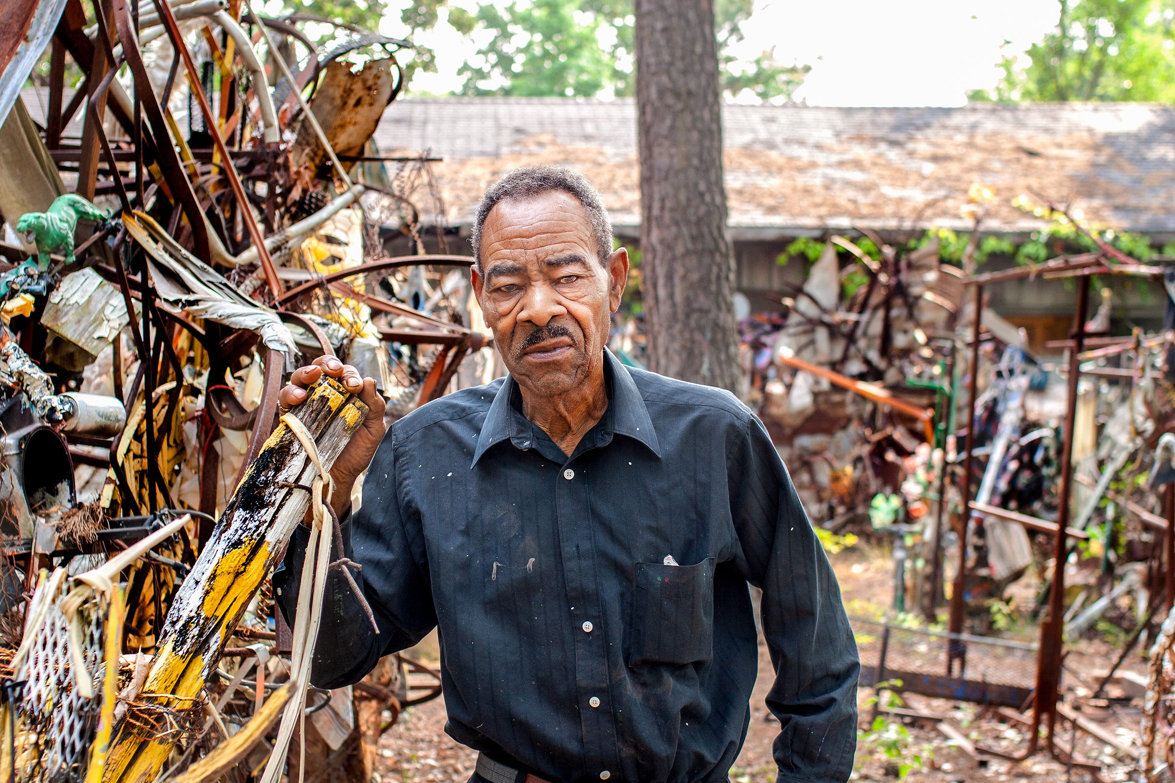 Thornton Dial stands in his yard in a dark shirt, one hand resting on a painted wooden strut, looking to one side. Around and behind him the yard is filled with his outdoor assemblages -- rusted frames, sheet metal, painted scrap and hanging cloth -- with the roof of a low building and trees beyond.