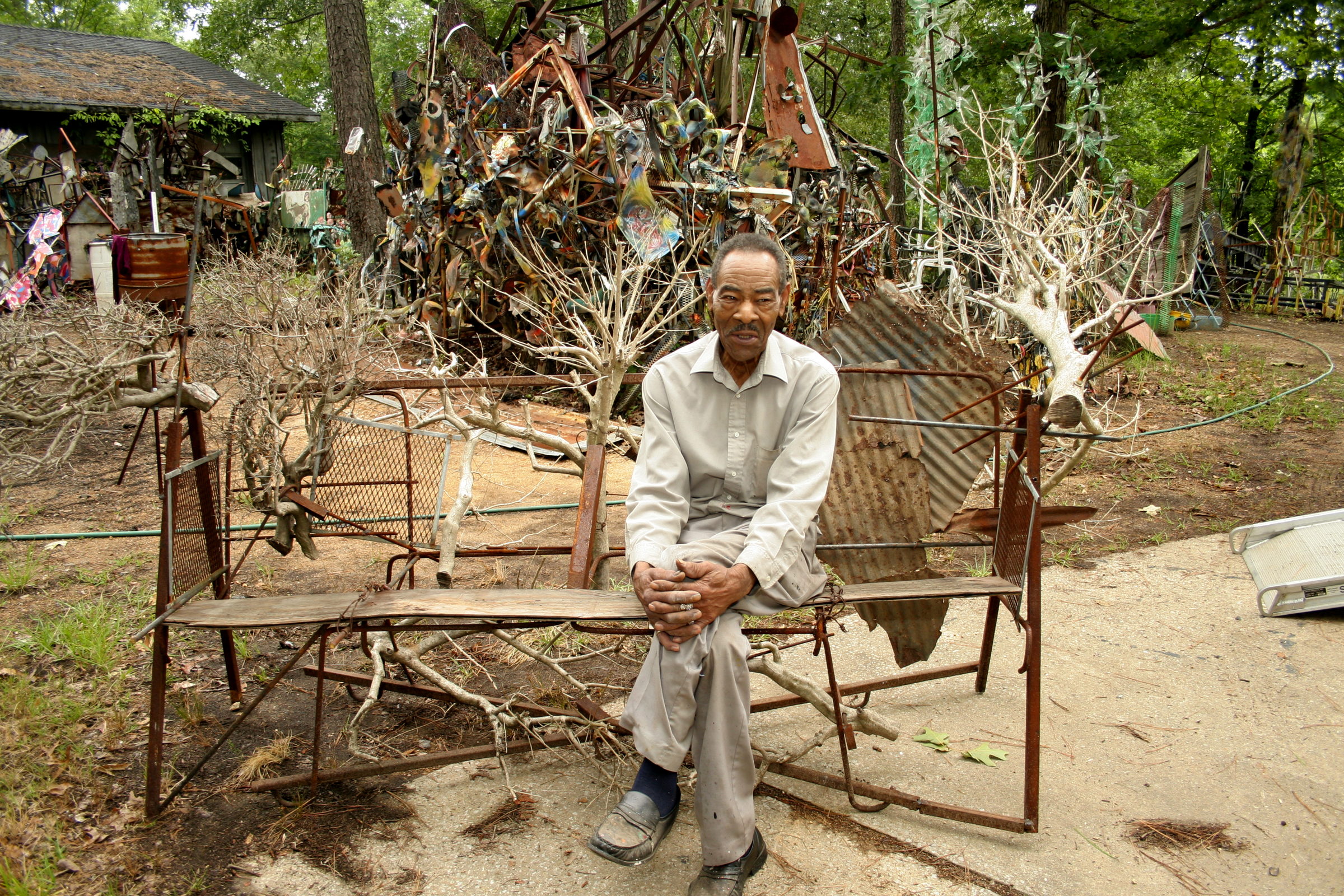 Thornton Dial sits on a bench made of welded rod and salvaged wood in the yard beside his house, hands folded, facing the camera. Behind him a tall assemblage of rusted metal and painted scrap rises among the trees, with bare-branched sculptures standing at either side and a shed to the left. A photograph by Stephen Pitkin.