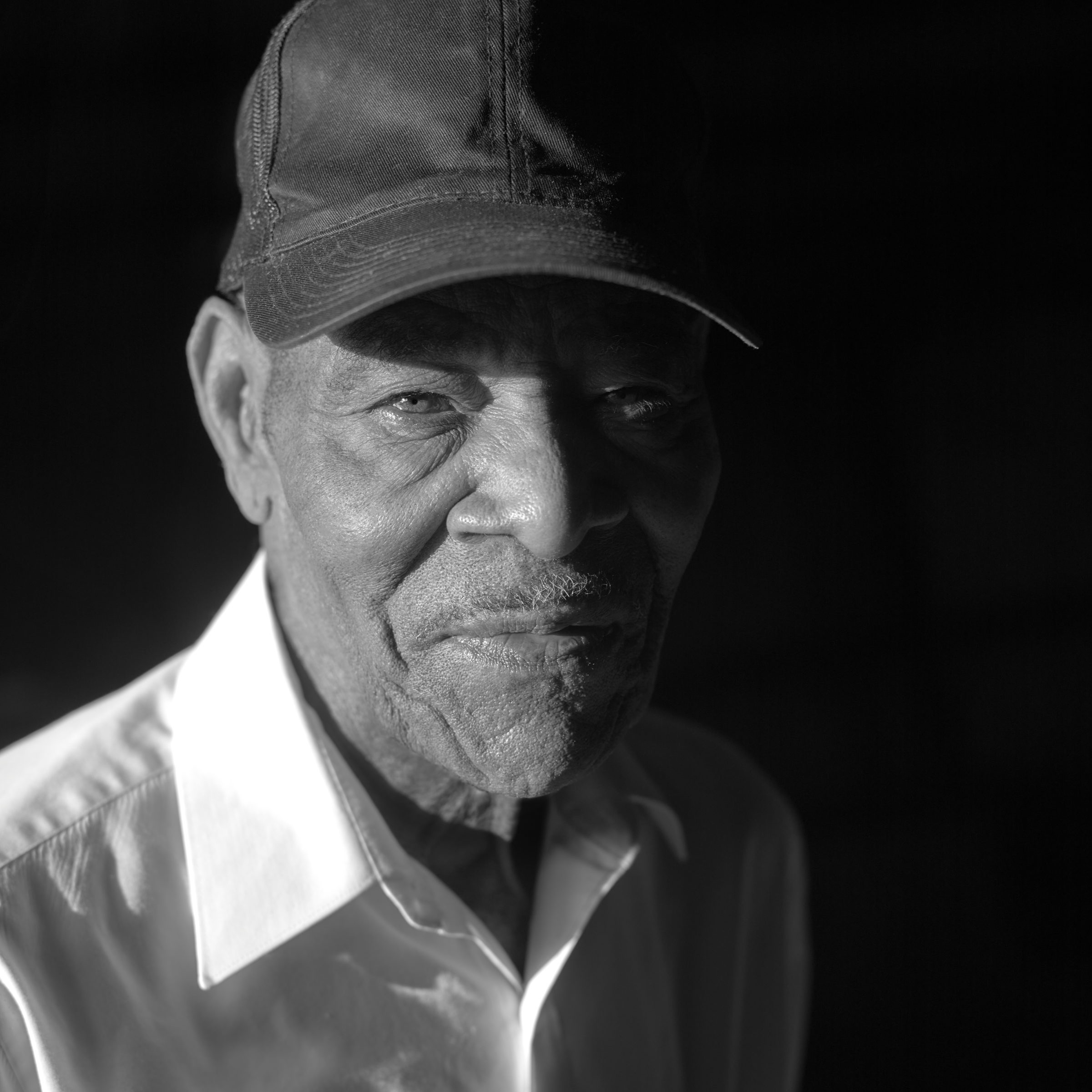 A close black-and-white portrait of Thornton Dial in later life against a black ground. He wears a dark work cap and an open-collared white shirt, his face lit from one side, looking just past the camera. A photograph by Stephen Pitkin.