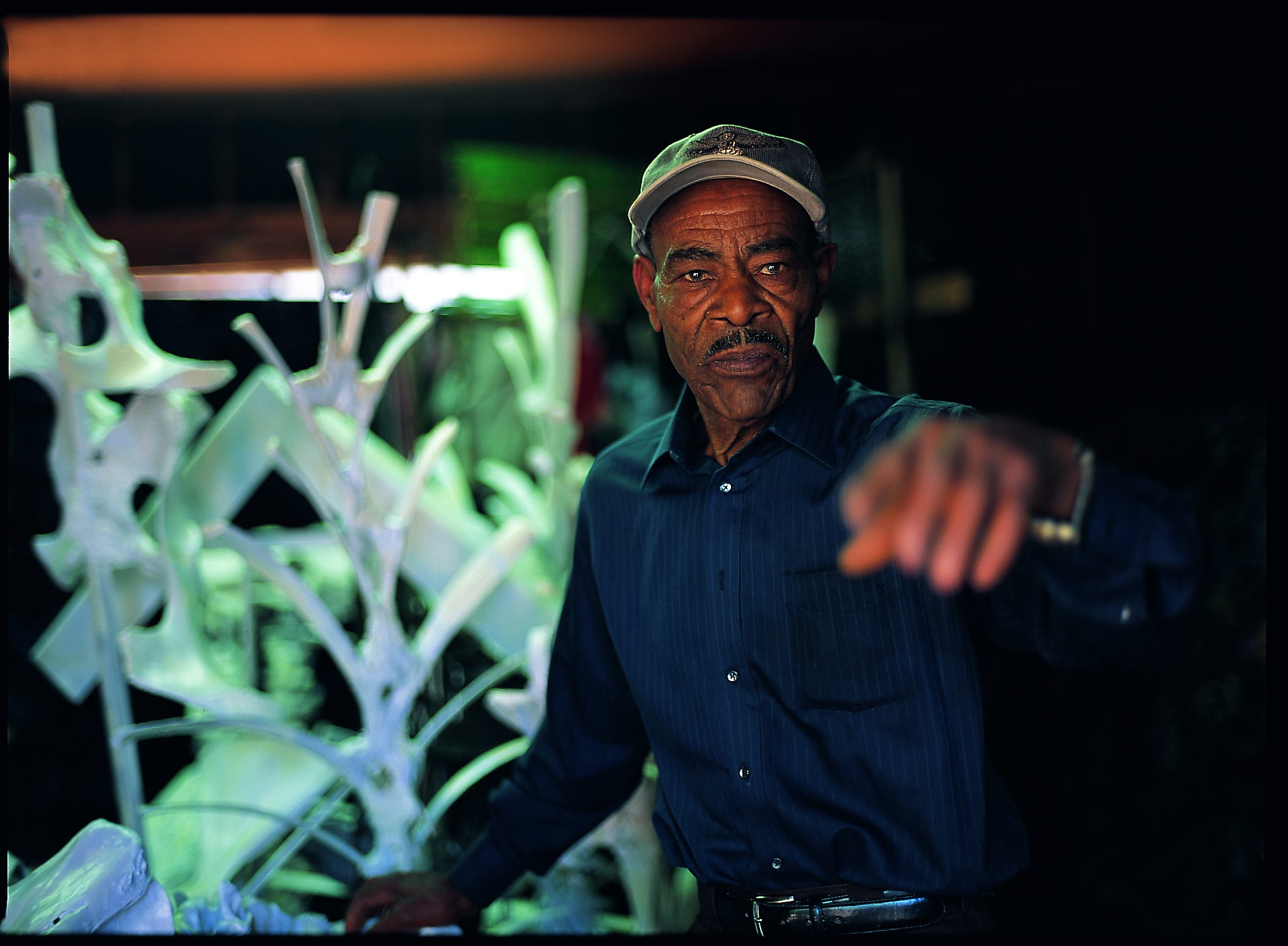 Thornton Dial in a dark shirt and pale cap in his workshop at night, pointing directly out of the frame toward the viewer. Beside him, lit green, stands a white-painted sculpture of branching bones and antler-like forms. A photograph by Stephen Pitkin.