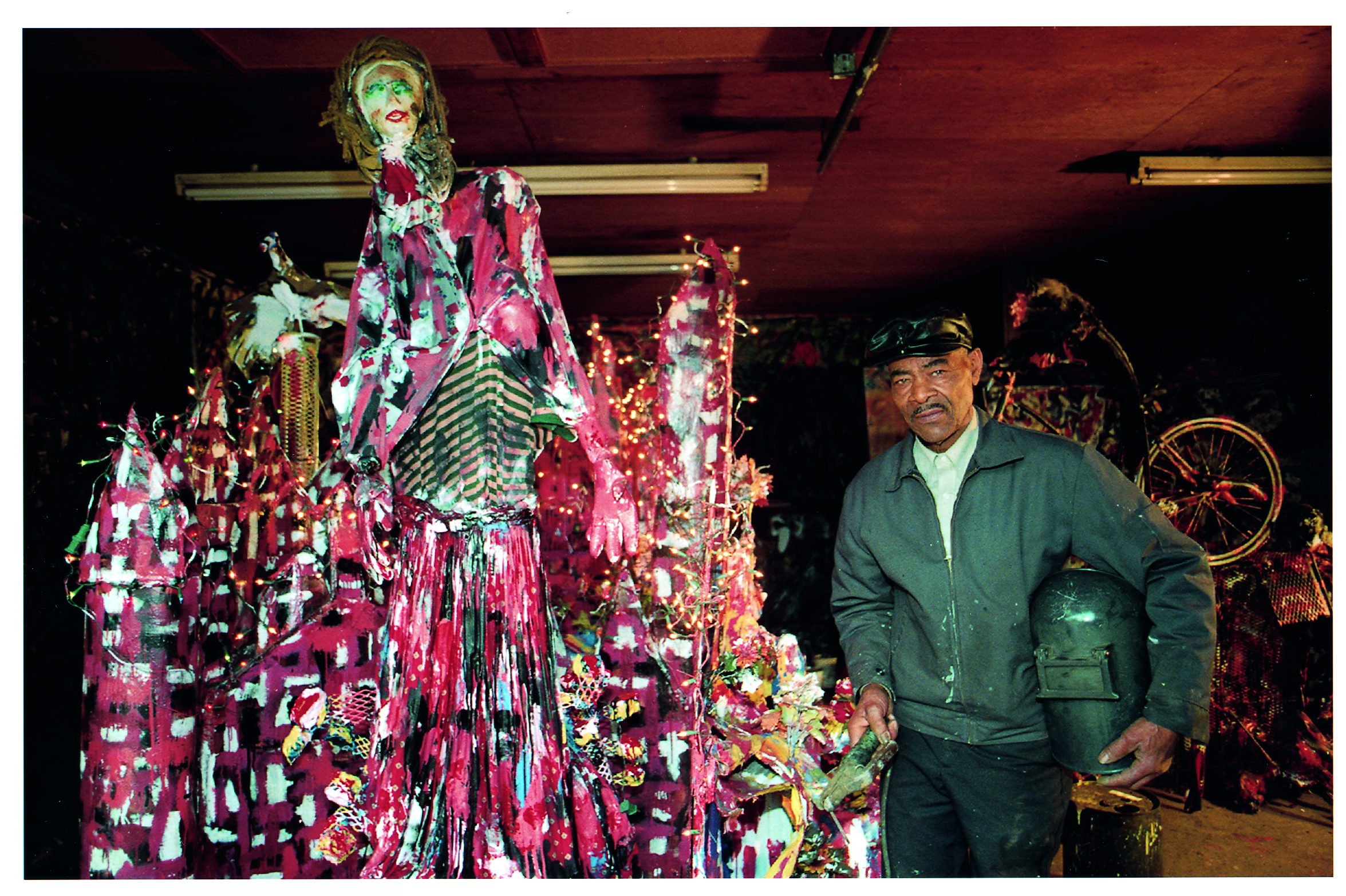Thornton Dial in work jacket and cap stands at the right of his lit workshop, holding a welding helmet under one arm. Filling the rest of the frame is a tall figure of a woman built from painted cloth in vivid pink and white, her arms outstretched over a skyline of jagged painted panels strung with fairy lights. A photograph by Stephen Pitkin.