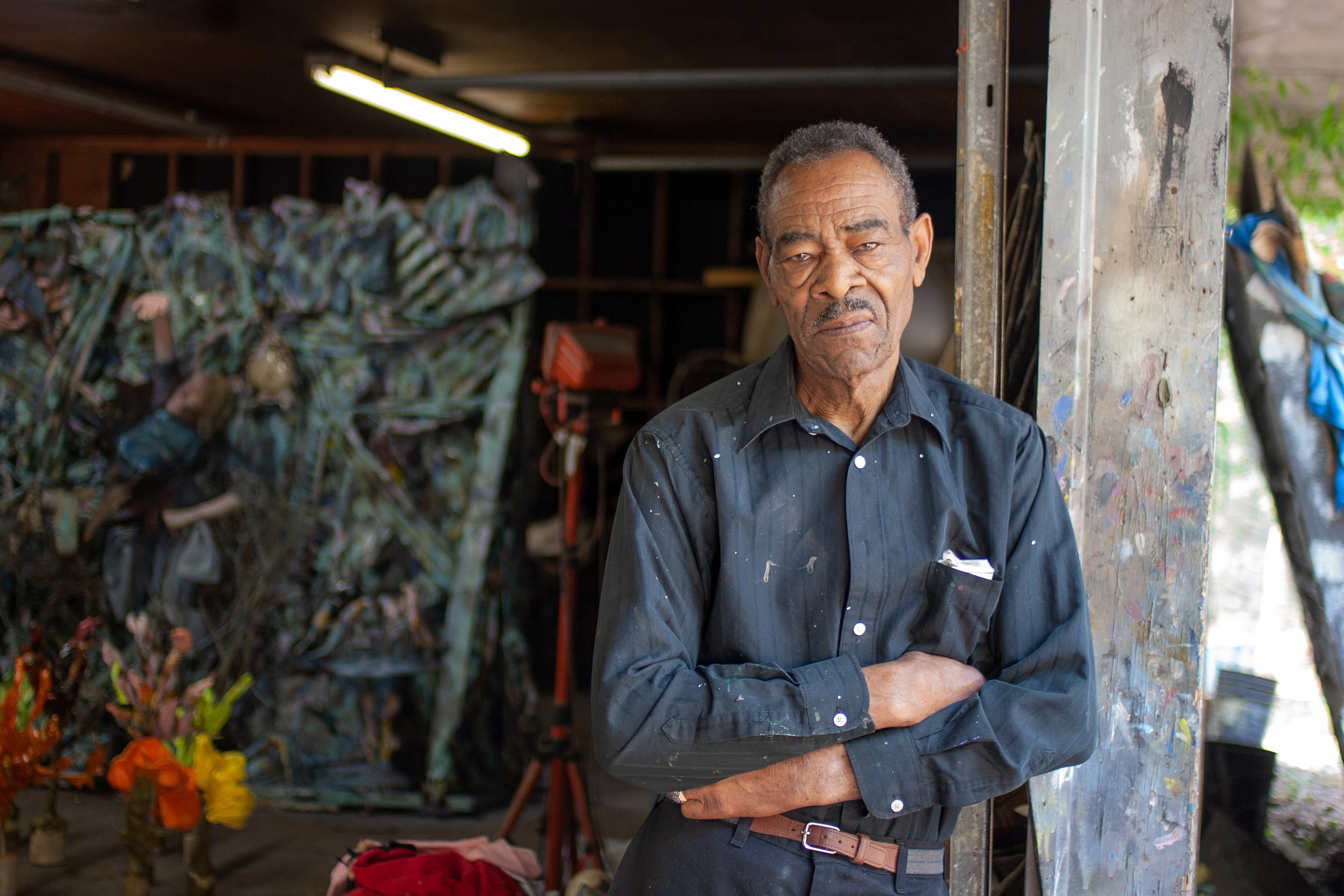 Thornton Dial stands with his arms folded at the open front of his studio shed, a paint-crusted post at his shoulder. Behind him a large blue-green assemblage leans against the wall and a work light stands on a red tripod. A photograph by Jerry Siegel.