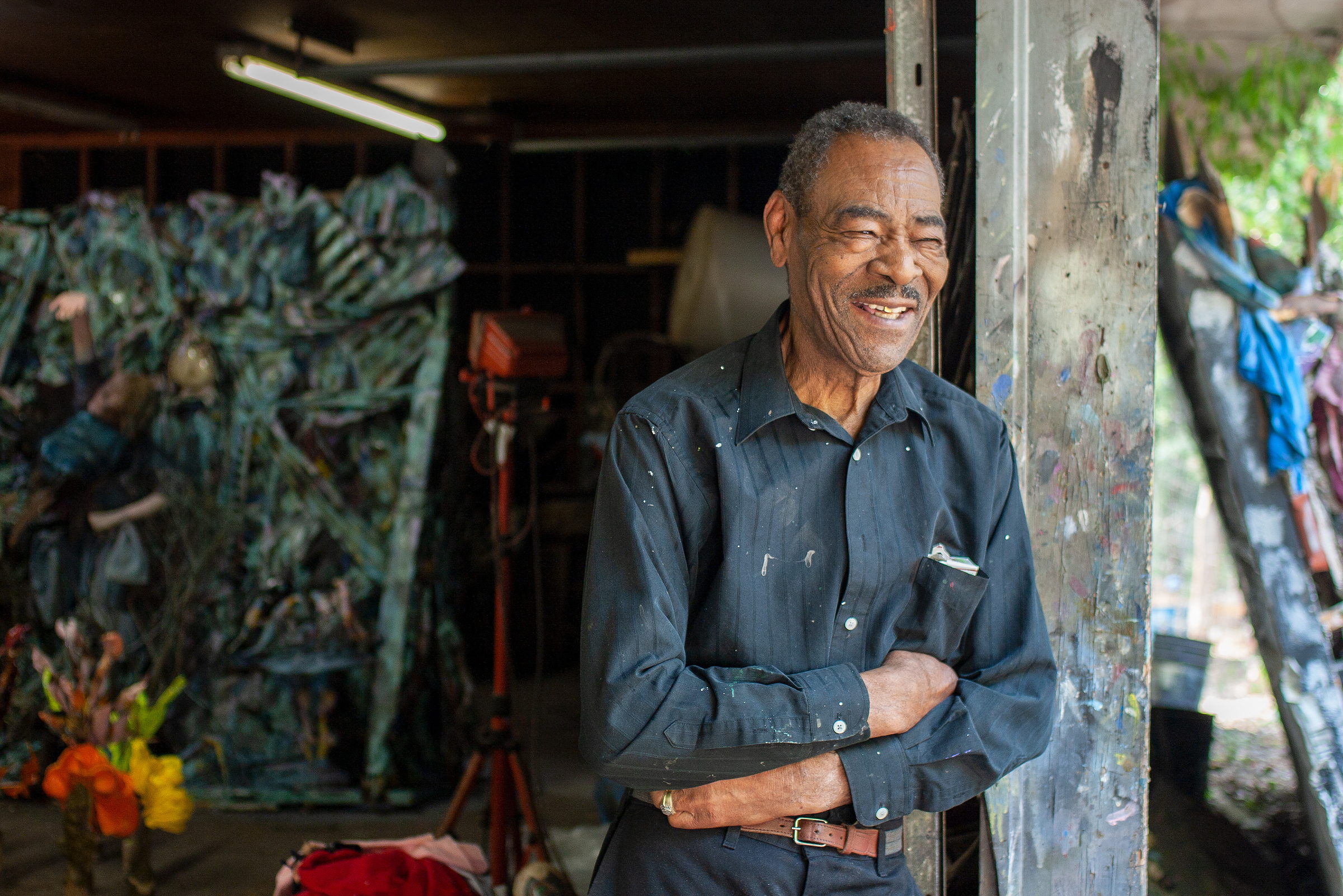 Thornton Dial laughing, head back, arms folded, leaning against the paint-covered post at the mouth of his studio. A large blue-green assemblage stands behind him in the shade of the shed. A photograph by Jerry Siegel.
