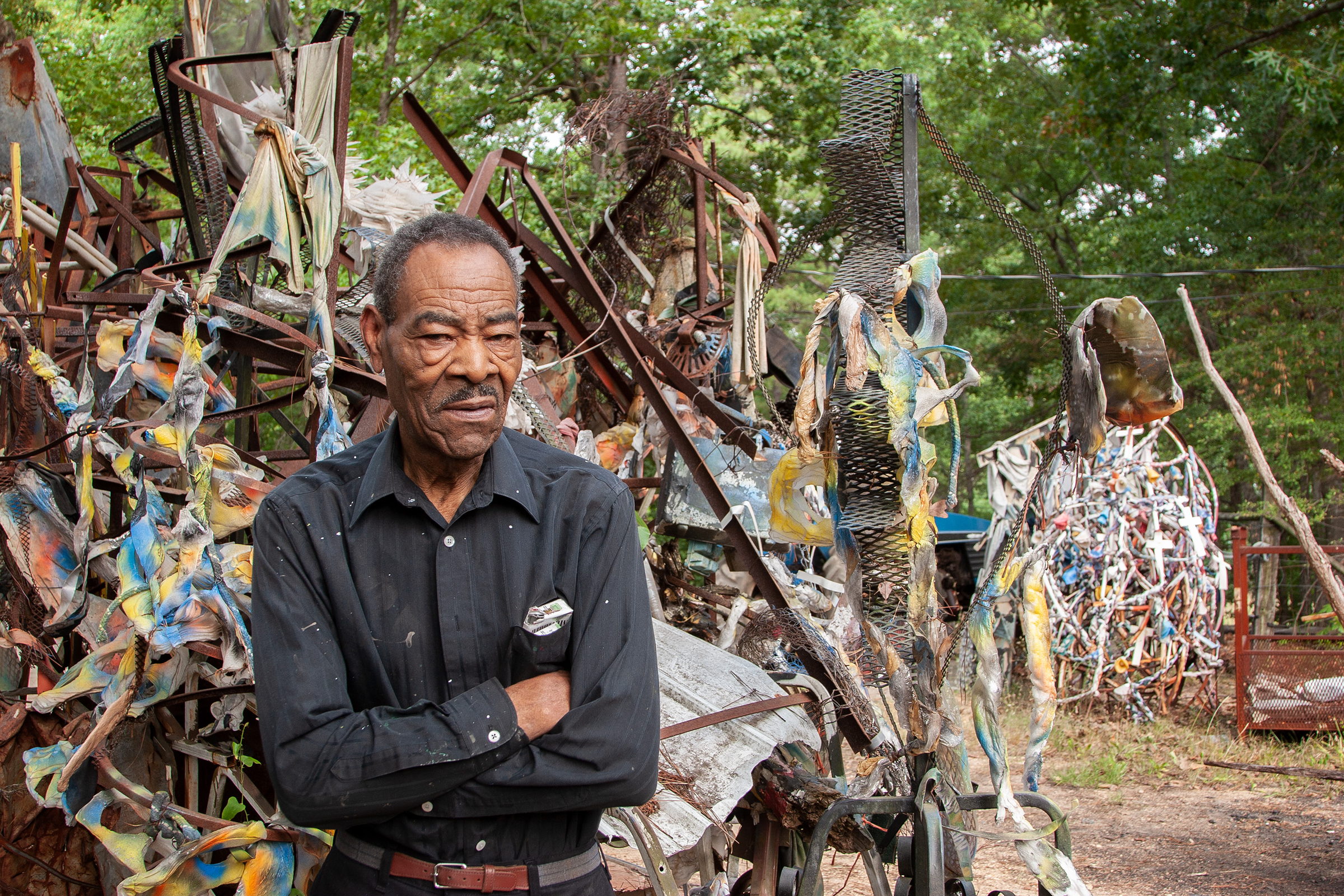Thornton Dial stands with arms folded in his yard, surrounded on all sides by his outdoor work: rusted steel frames hung with painted cloth in blue, yellow and white, lengths of wire mesh, and a tangled sphere of white-painted scrap to the right. A photograph by Jerry Siegel.