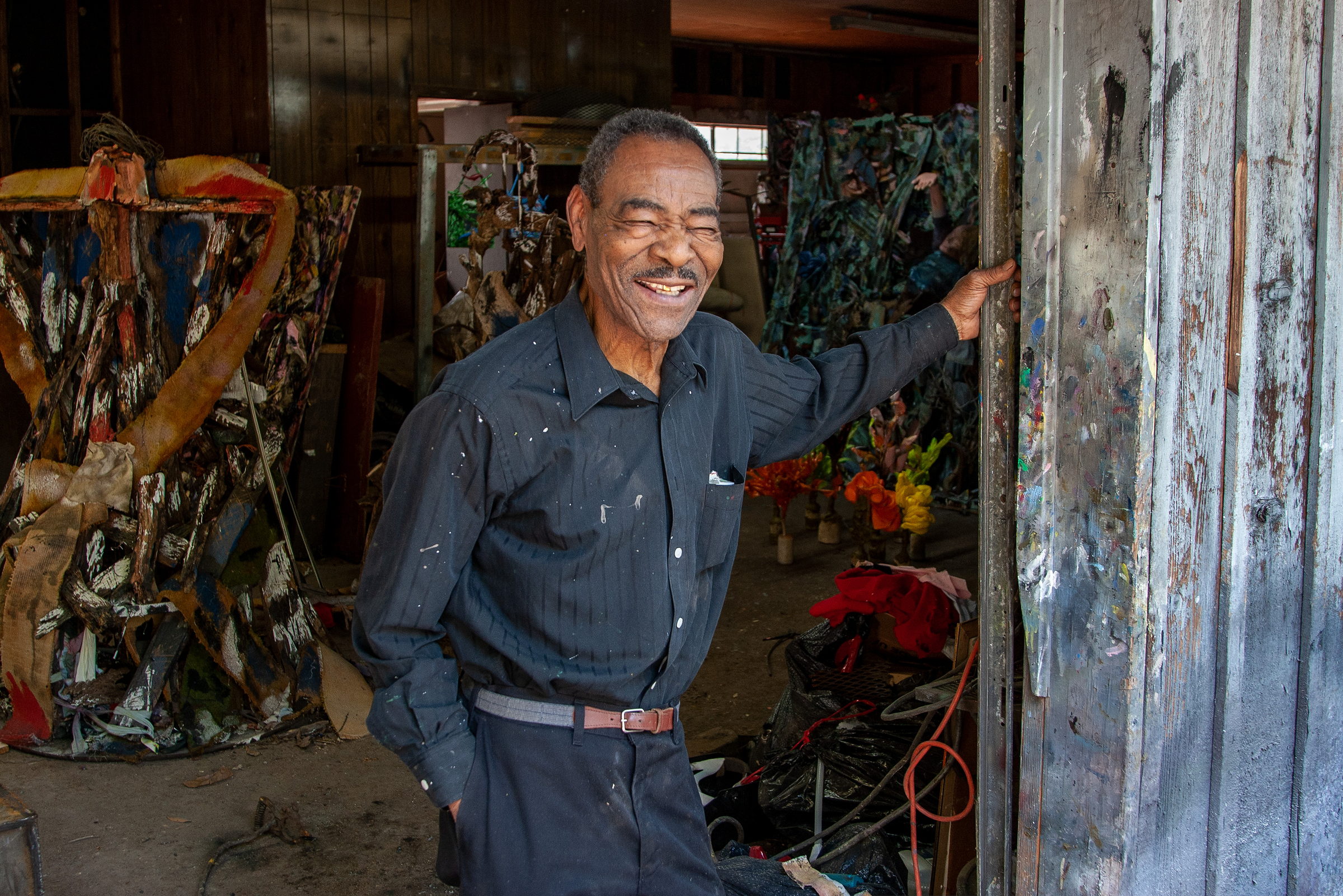 Thornton Dial grinning broadly, one hand raised against the studio door frame, the other in his pocket. To the left a tall assemblage of torn and painted material stands on the floor; deeper in the shed more works are stacked against the wall. A photograph by Jerry Siegel.