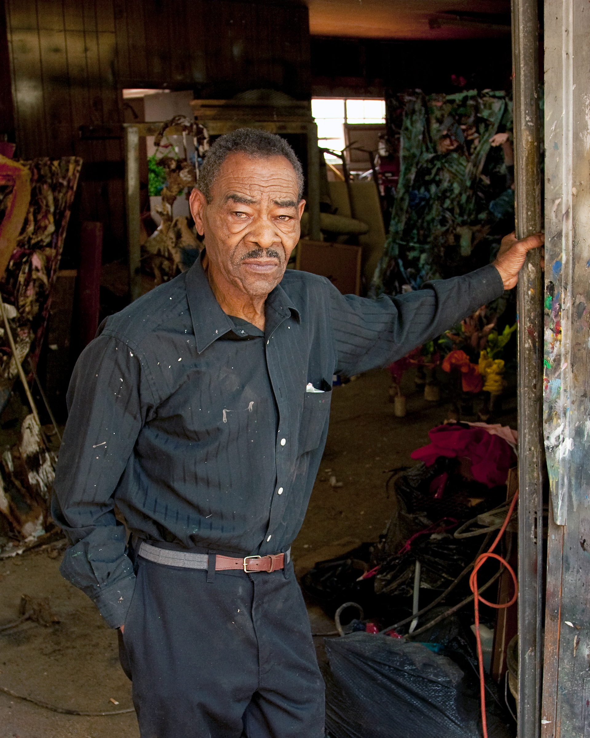 Thornton Dial stands in the doorway of his studio in a paint-spattered dark shirt, one hand on the frame, looking steadily at the camera. Behind him the interior is stacked with works in progress and bundled materials; the door post beside him is crusted with years of paint. A photograph by Jerry Siegel.