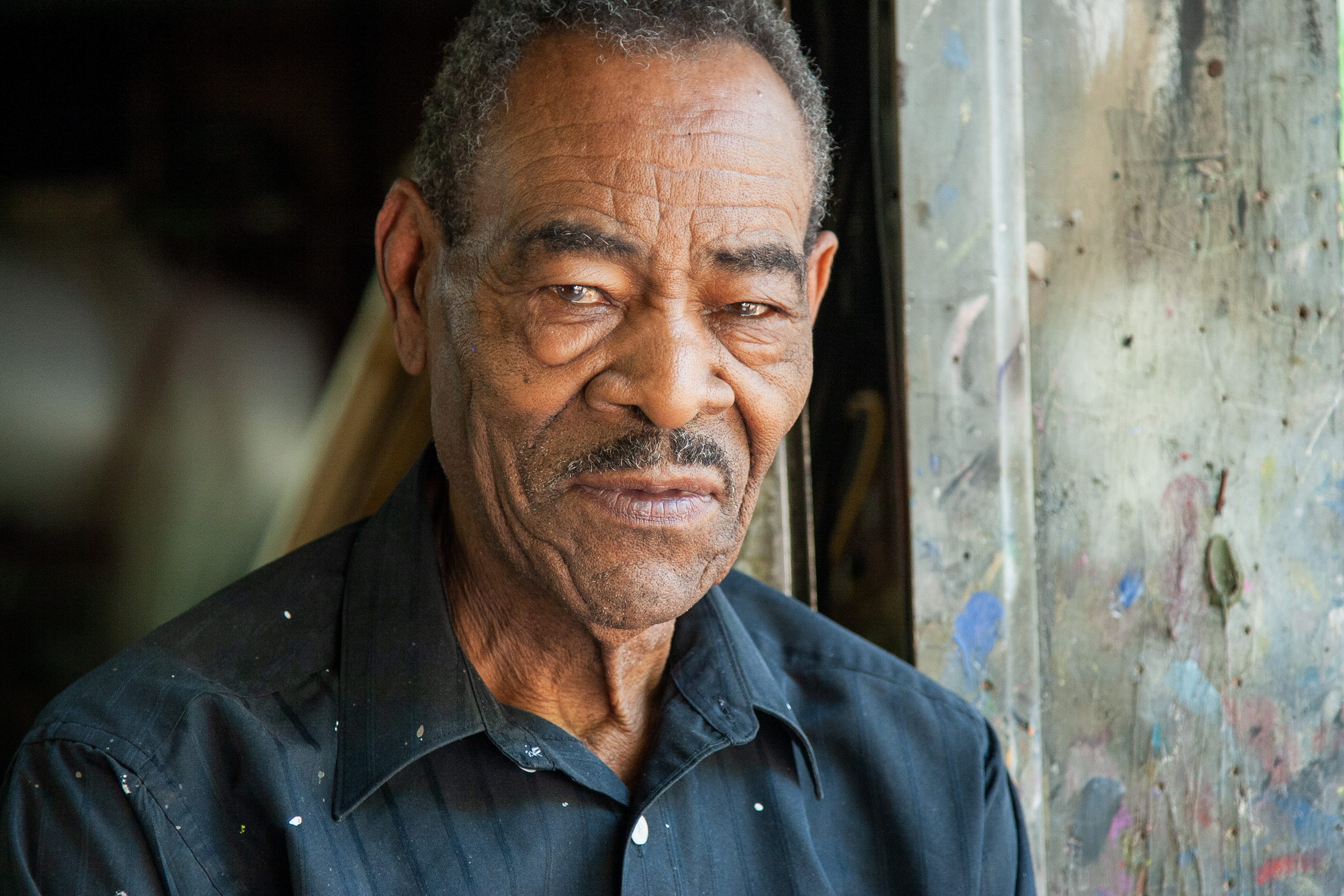 A close portrait of Thornton Dial in a dark shirt flecked with white paint, turned slightly and looking past the camera, half-smiling. The paint-spattered studio door frame fills the right of the picture. A photograph by Jerry Siegel.