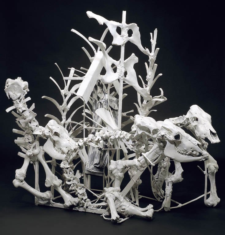 A freestanding sculpture, photographed against black, built from animal bones and found metal and painted entirely white. Long leg bones and vertebrae are wired into a framework that rises to the left and right, with two large animal skulls turned outward at the front, a smaller one at the upper left, and a chair-like structure held in the middle of it all.