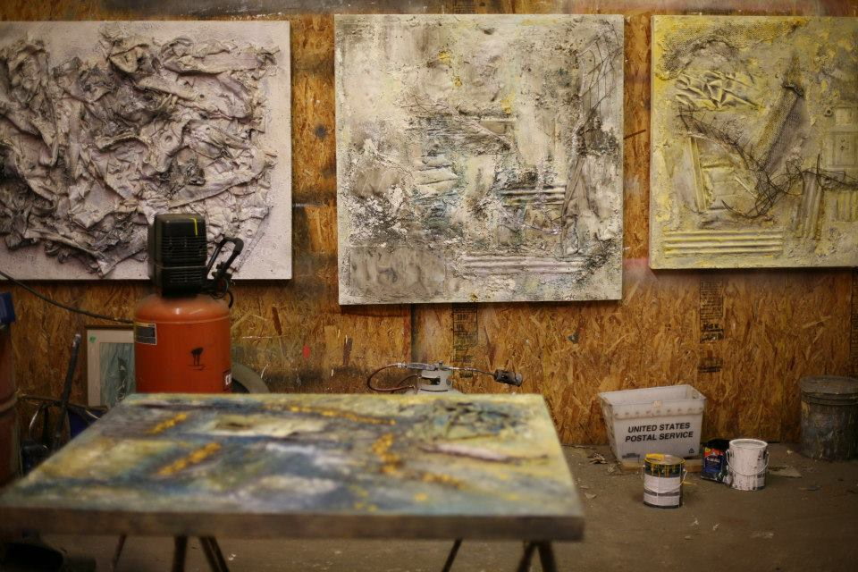 Three large pale assemblages hang on the plywood wall of the studio -- one white and heavily encrusted, one grey and green, one yellow -- above a paint compressor and a stack of paint cans. In the foreground a blue and yellow panel lies flat on a trestle, still being worked on.