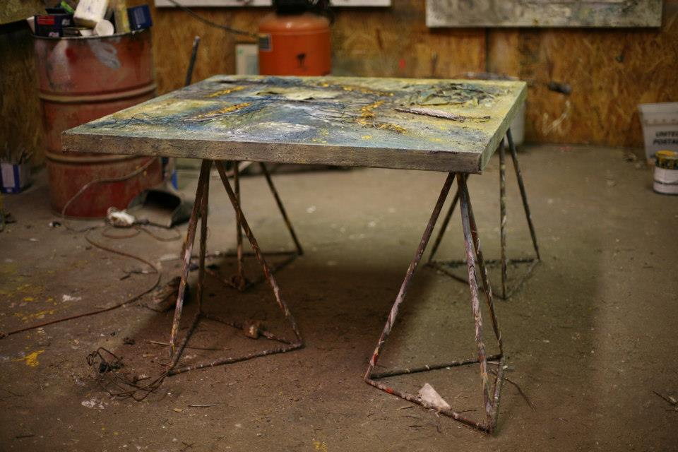 A blue and gold panel lies flat on two welded metal trestles in the middle of the studio floor, with fresh material still heaped on its surface. Around it are a rusted oil drum of brushes, scattered paint cans, and a concrete floor marked with years of spill.