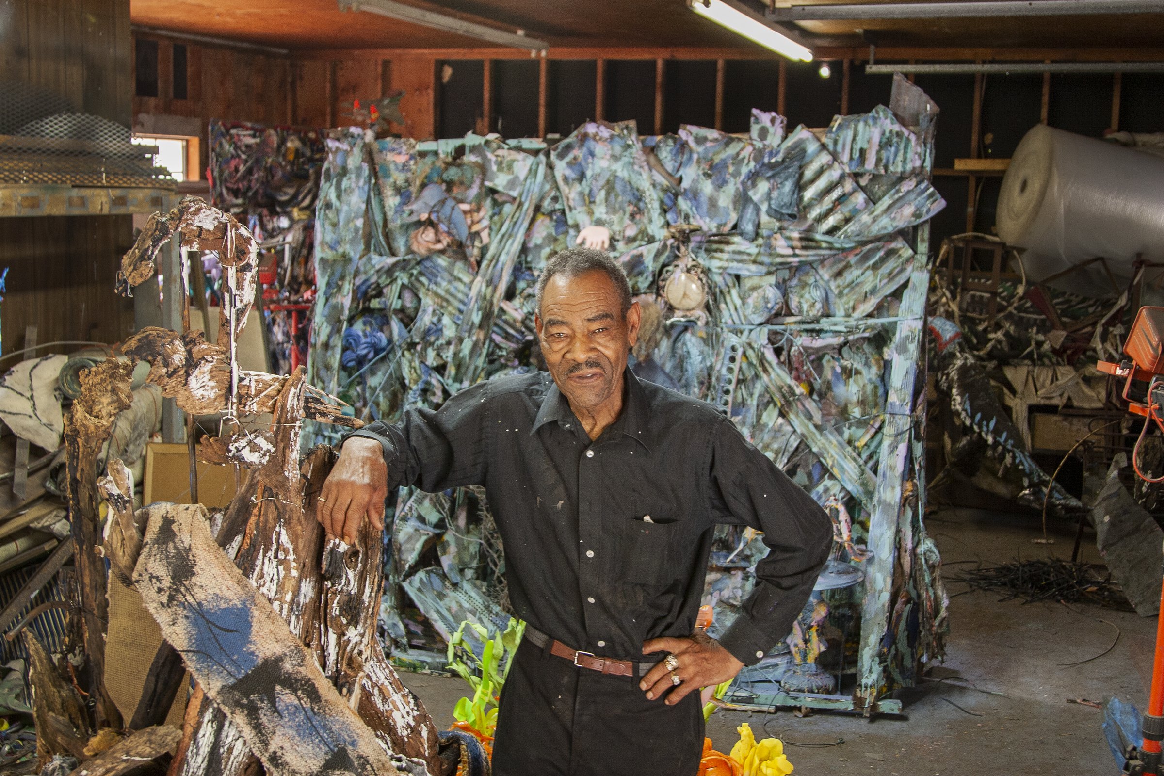 Thornton Dial standing in his studio, one hand on his hip and the other resting on a tall weathered piece of painted driftwood. Filling the wall behind him is a huge assemblage of torn and folded material painted blue-green, and around him the floor is stacked with more work, rolls of material and artificial flowers.