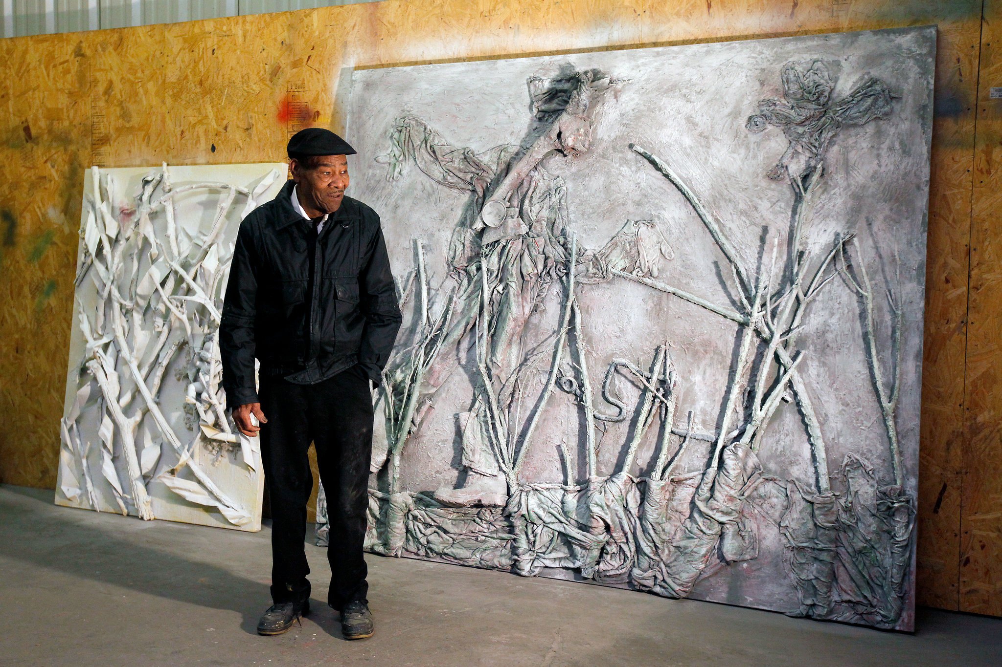 Thornton Dial stands in his studio in a dark jacket and flat cap, hands at his sides, looking to one side. Behind him lean two large relief works painted entirely white and grey: a smaller one of tangled branches and cloth, and a wall-sized panel in which draped figures and long bare branches rise out of the surface.