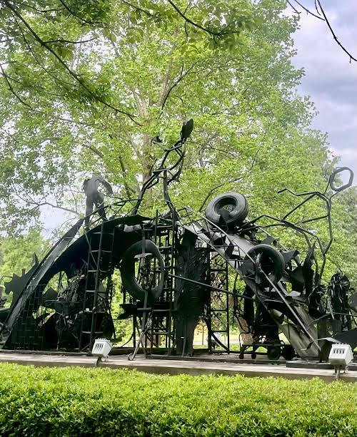 An outdoor sculpture in a public park: a wide arch of welded black steel, perhaps two storeys high, built from salvaged wheels, tyres, sheet metal, ladders and curling rods, standing on a paved plaza behind a clipped hedge with mature trees rising behind it.